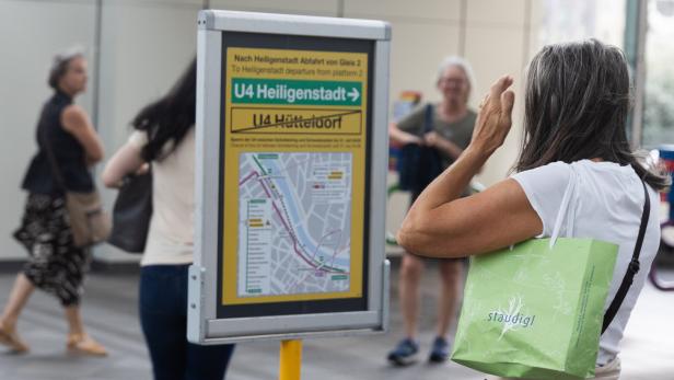 Eine Frau mit grüner Einkaufstasche betrachtet einen U-Bahn-Plan an einer Station, während andere Personen vorbeigehen.