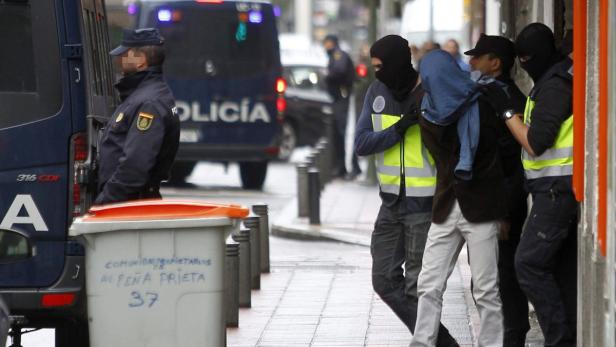 Polizisten führen eine verhaftete Person ab, deren Gesicht verdeckt ist.