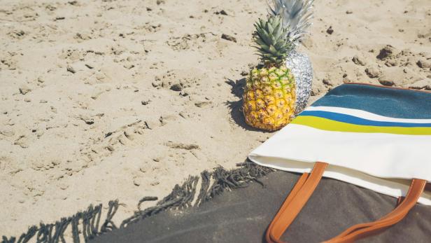 Eine Ananas und eine silberne Ananas-Skulptur liegen neben einer Strandtasche im Sand.