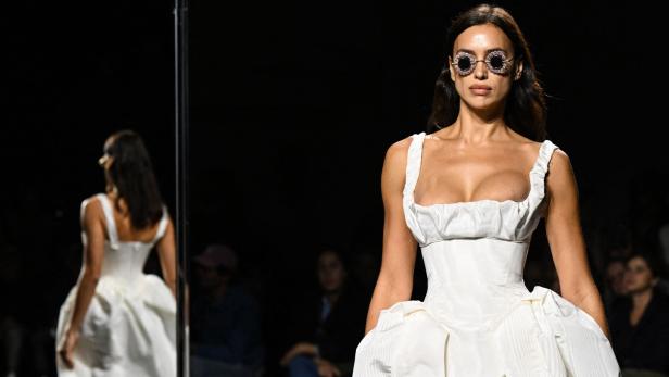 Irina Shayk trägt ein weißes Kleid und runde Sonnenbrille auf dem Laufsteg.
