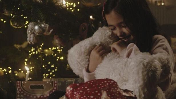 Ein Mädchen umarmt einen Teddybären vor einem Weihnachtsbaum mit Geschenken.