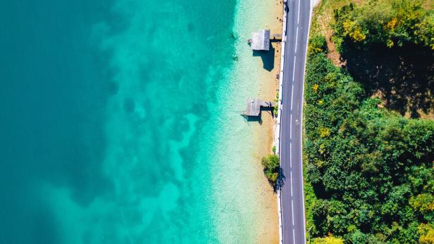 Eine Drohnenaufnahme vom Attersee: Eine Wiese mit Bäumen, dann eine Straße am Ufer und daran grenzt der klare Attersee in verschiedenen Blautönen.