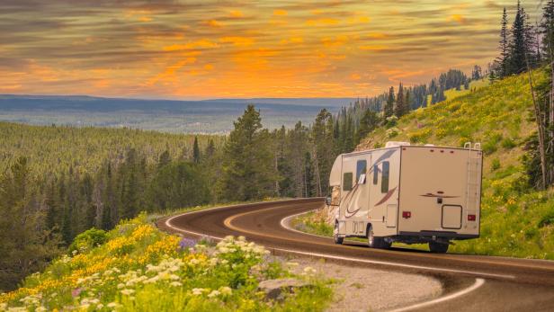 Wohnmobil fährt eine kurvige Straße entlang, im Hintergrund Wiesen und Wald bei Sonnenuntergang