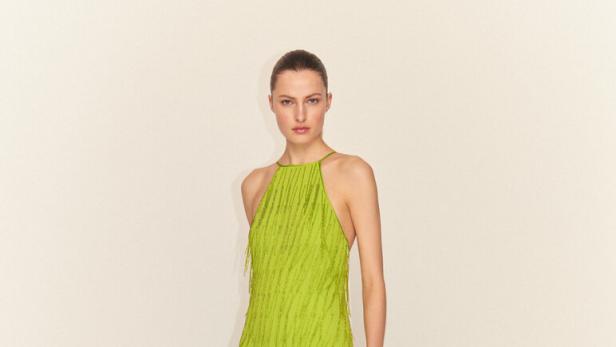 Eine Frau in einem langen, hellgrünen Fransenkleid vor einem neutralen Hintergrund.