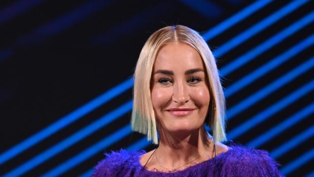 Sarah Connor mit blondem Bob-Haarschnitt und lila Oberteil vor einem blau gestreiften Hintergrund.