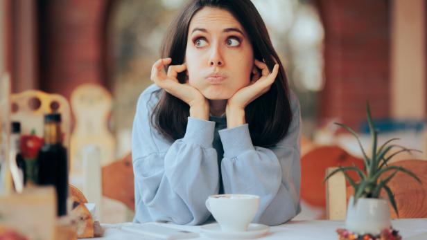 Eine gelangweilte Frau sitzt an einem Tisch mit einer Tasse Kaffee.