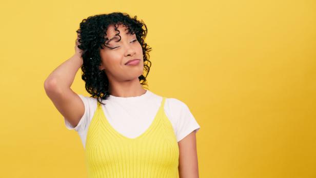Eine Frau mit lockigem Haar kratzt sich nachdenklich am Kopf vor einem gelben Hintergrund.