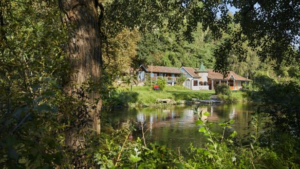 Grün-braunes Wasser, Bäume, eine Liegewiese und im Hintergrund ein Holzgebäude, das einst das Flussbad beherbergte
