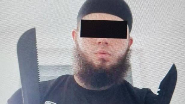 Ein Mann mit Bart und Mütze hält zwei große Messer vor der Brust.