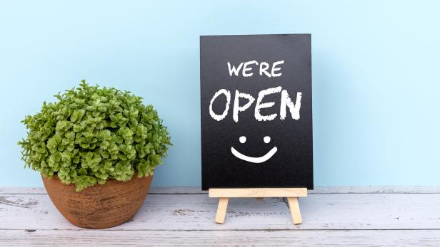 Zu sehen ist eine weiße Tafel neben einer Pflanze auf der steht: "We are open" mit einem Smiley