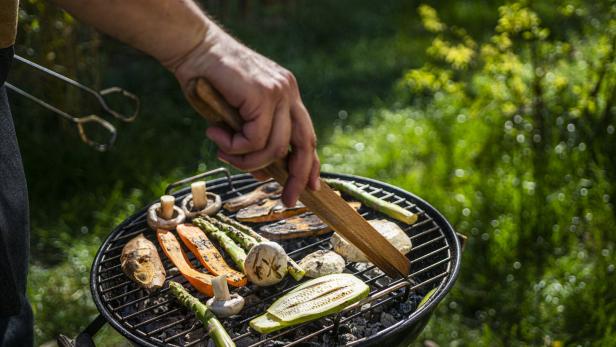 Kleiner Griller, auf dem Rost liegen Zucchini, Champignons, Spargel und Melanzani, ein Mann wendet das Gemüse mit einer Grillzange