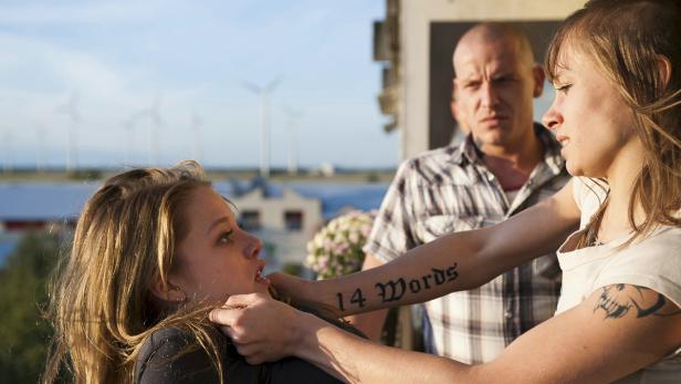 Eine Frau wird von einer anderen Frau angegriffen, während ein Mann zusieht. Die Angreiferin hat ein Tattoo mit der Aufschrift „14 Words“.