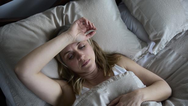 Eine Frau liegt im Bett und hält die Hand auf die Stirn.