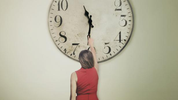 Eine Frau in einem roten Kleid versucht, die Zeiger einer großen Uhr zu verstellen.