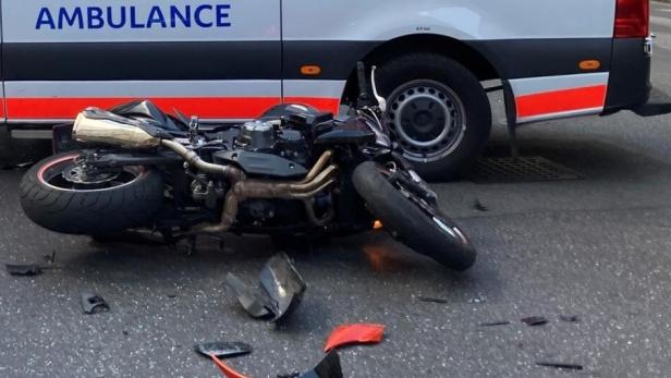 Umgekippte Motorrad-Unfallstelle vor einem Krankenwagen mit Trümmern auf der Straße.