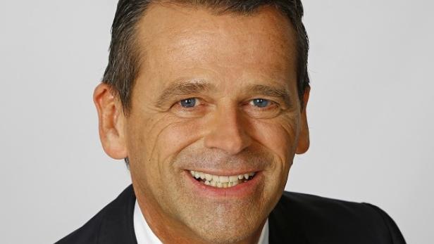 Ein lächelnder Mann im Anzug vor einem hellen Hintergrund.