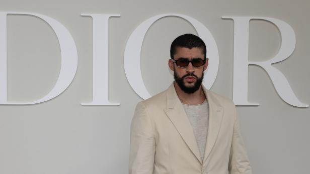 Ein Mann mit Sonnenbrille und hellem Anzug steht vor einem Dior-Hintergrund.