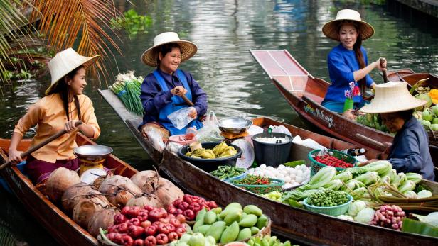 Frauen in traditionellen thailändischen Booten verkaufen frisches Gemüse und Obst, während sie auf einem malerischen Fluss fahren