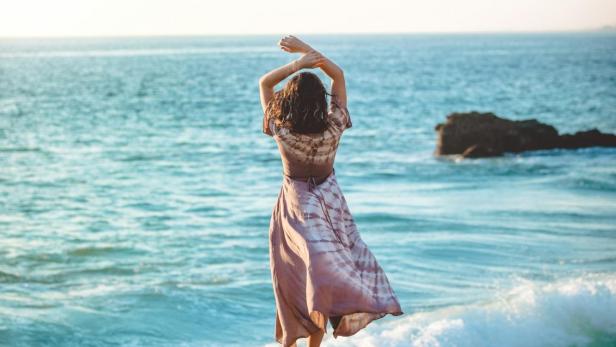 Eine Frau in einem Kleid steht mit erhobenen Armen an der Küste.