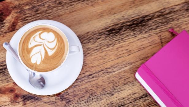 Ein Cappuccino mit Latte Art und ein pinkfarbenes Notizbuch auf einem Holztisch.