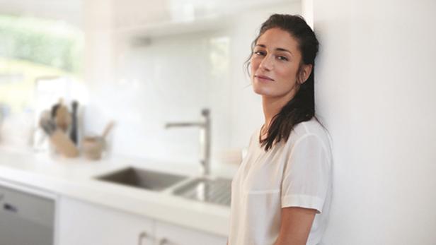Eine Frau lehnt an einer Wand in einer hellen Küche.