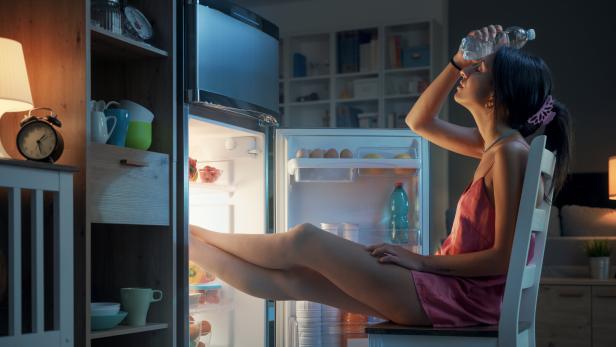 Eine Frau sitzt unter der Hitze leidend vor dem offenen Kühlschrank.