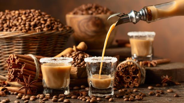 Ein Getränk wird in ein Glas mit Zuckerrand gegossen, umgeben von Kaffeebohnen und Zimtstangen.