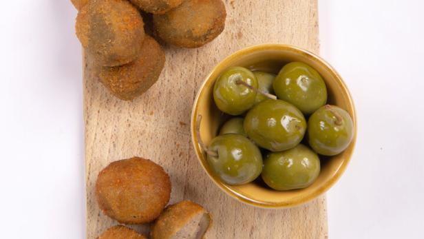 Panierte Oliven und ein Schälchen mit grünen Oliven auf einem Holzbrett.