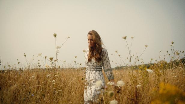 Eine Frau mit langem Haar geht durch ein Feld mit hohem Gras und Wildblumen.