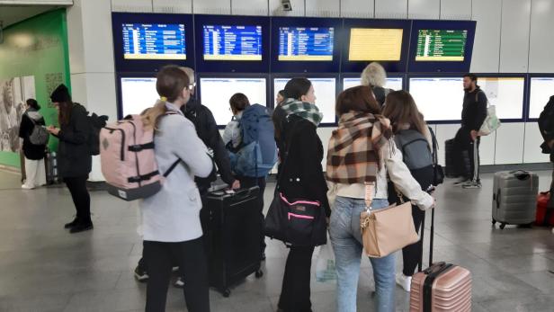 Reisende warten mit Gepäck auf einem Bahnhof vor Anzeigetafeln.