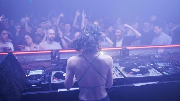 Eine junge Frau legt als DJ Platten in einem Techno-Club auf – von hinten fotografiert. Vor ihr jubeln Menschen.