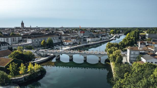 Eine Brücke über dem Fluss Charente. Am Ufer stehen Teile der Stadtmauer, ein großes Gebäude des Cognac-Herstellers Hennessey, im Hintergrund erhebt sich die Kirche.