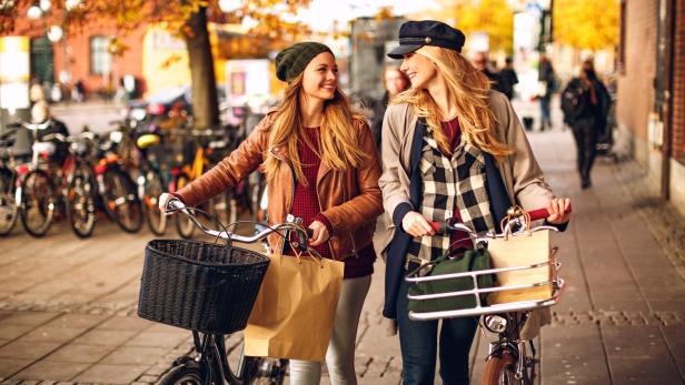 Zwei junge Frauen schieben ihre Fahrräder auf einem Bürgersteig im Herbst.
