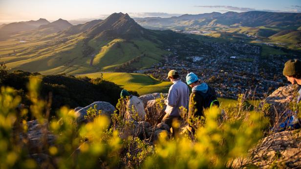 Eine Gruppe von Wanderern genießt die Aussicht auf eine Stadt und grüne Hügel im goldenen Abendlicht.