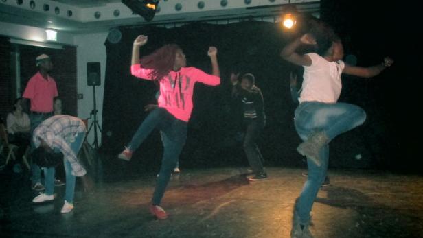 Eine Gruppe junger Leute tanzt auf einer Bühne vor einem schwarzen Hintergrund.