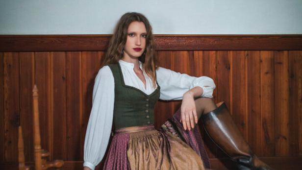 Eine Frau in einem Lena Hoschek Dirndl sitzt auf einer Holzbank neben einem Spinnrad.