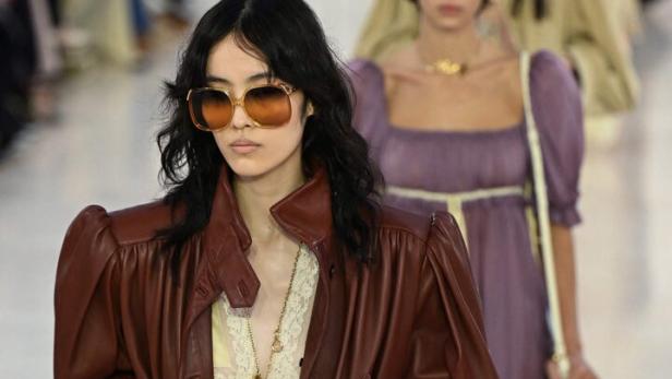 Eine Frau mit Sonnenbrille und brauner Lederjacke läuft auf einer Modenschau.
