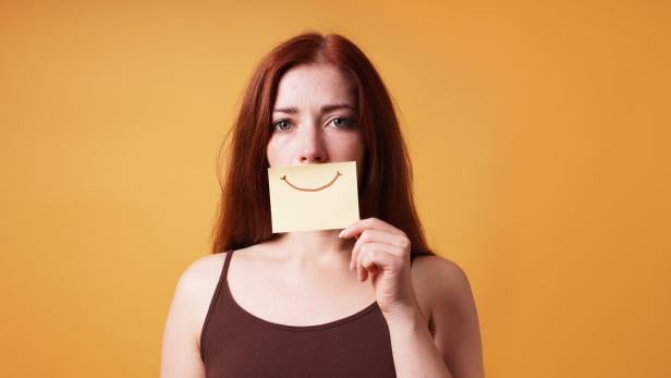 Eine Frau hält sich ein Post-It mit einem aufgemalten Lächeln vor ihren Mund..