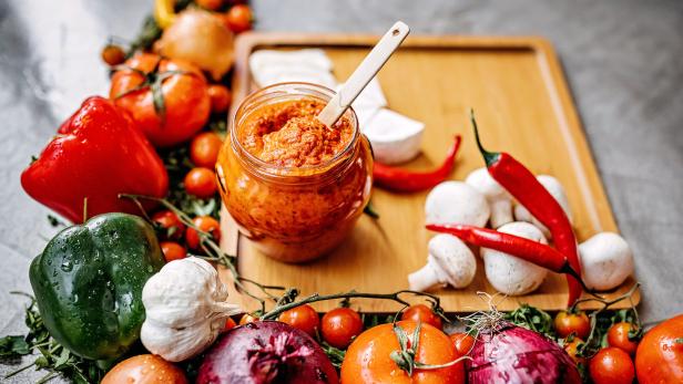 Ein Glas Ajvar umgeben von frischem Gemüse wie Paprika, Tomaten und Zwiebeln.