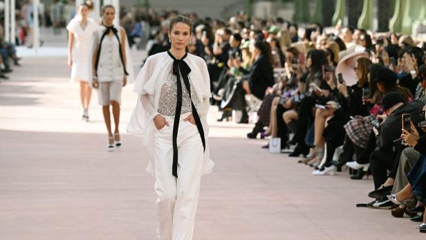 Ein Model geht über den Laufsteg, in weißen Hosen und einem weißen Cape.