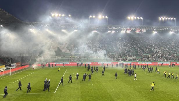 Polizeibeamte betreten ein Fußballfeld, das mit Rauch gefüllt ist, während Fans auf den Tribünen sitzen.