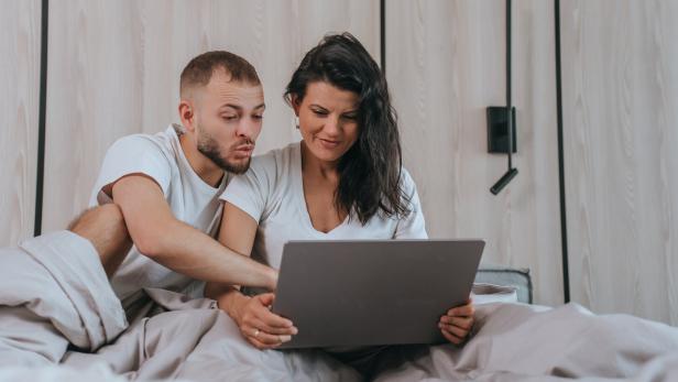 Ein Paar sitzt im Bett und schaut auf einen Laptop.