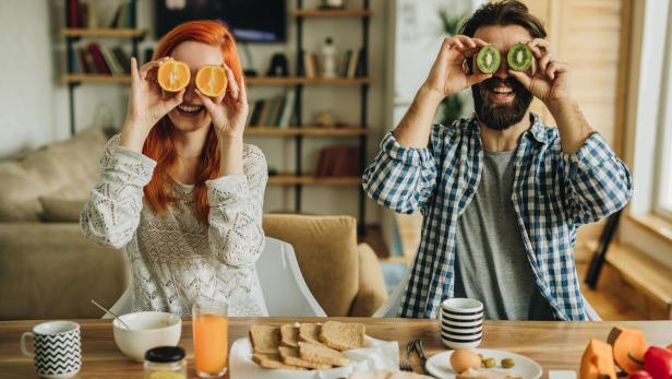 Ein Mann und eine Frau sitzen beim Frühstück und halten sich Orangen- und Kiwihälften vor die Augen.
