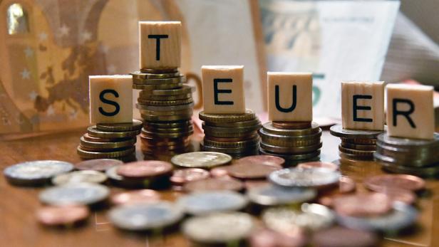 Holzbuchstaben auf Münzstapeln bilden das Wort „Steuer“, im Hintergrund sind Geldscheine zu sehen.