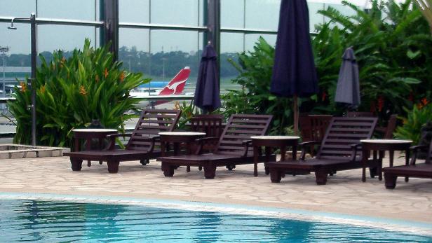 Liegestühle und Sonnenschirme stehen an einem Pool mit Blick auf einen Flughafen.