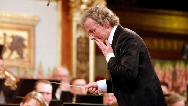 Ein Dirigent mit Brille und Anzug dirigiert ein Orchester.