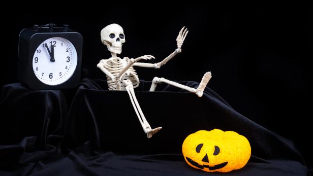 Ein Skelett sitzt neben einer leuchtenden Kürbis-Dekoration und einer Uhr, die fast Mitternacht zeigt.