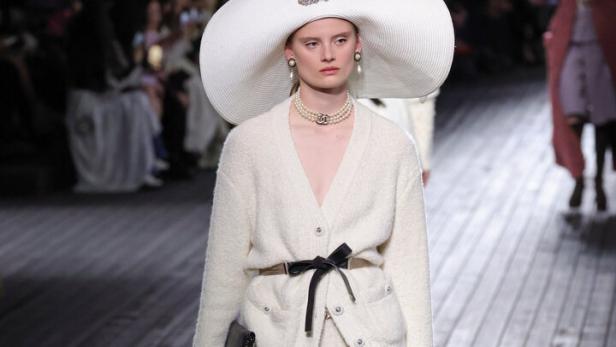 Ein Model mit übergroßem Hut und cremefarbenem Hosenanzug auf einem Laufsteg.