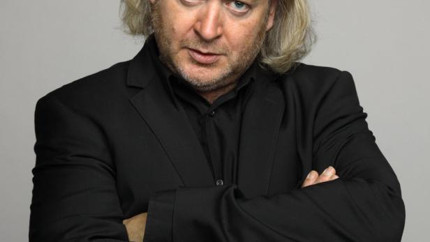 Ein Mann mit langen blonden Haaren und blauem Blick, im schwarzen Anzug, verschränkt die Arme vor der Brust.