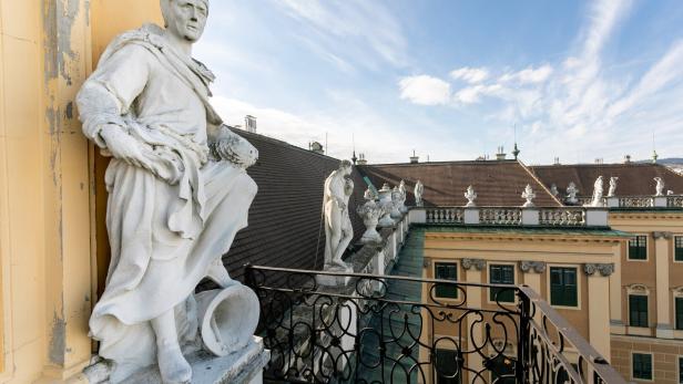 Steinfiguren schauen beim Balkon unterhalb der großen Uhr von oben auf den Ehrenhof des Schlosses Schönbrunn herab. Vor dem Balkon befindet sich ein Schmiedeisengitter, vor dem Dach eine steinerne Brüstung